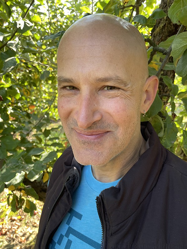 Jesse standing in front of a tree, smiling and wearing a black down vest over a blue t-shirt.