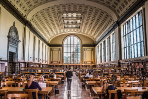 The North Reading Room of the UC Berkeley Library. It is a long room with a vaulted ceiling, many tables at which students are sitting, and an enormous window at the end.