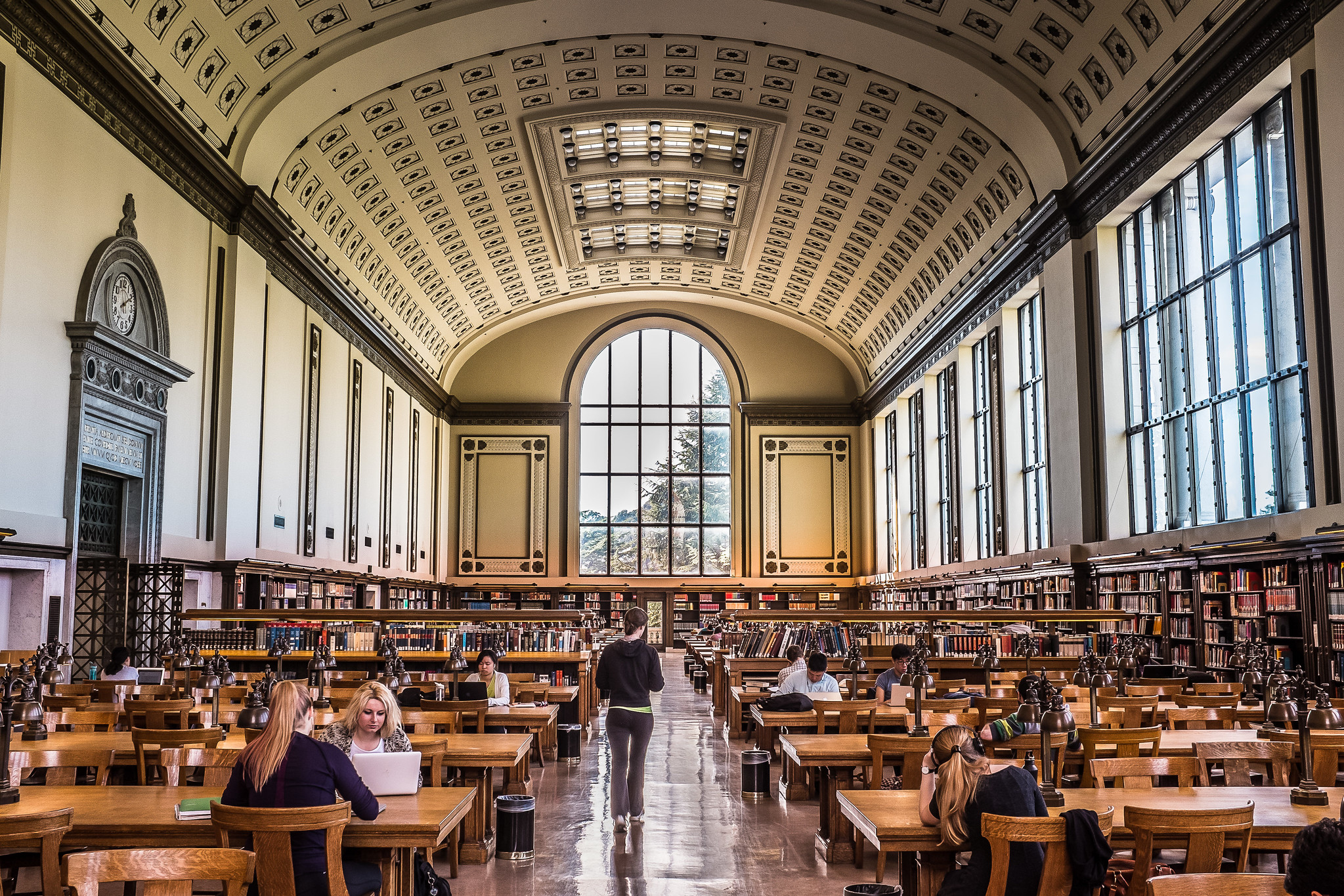 The North Reading Room of the UC Berkeley Library. It is a long room with a vaulted ceiling, many tables at which students are sitting, and an enormous window at the end.
