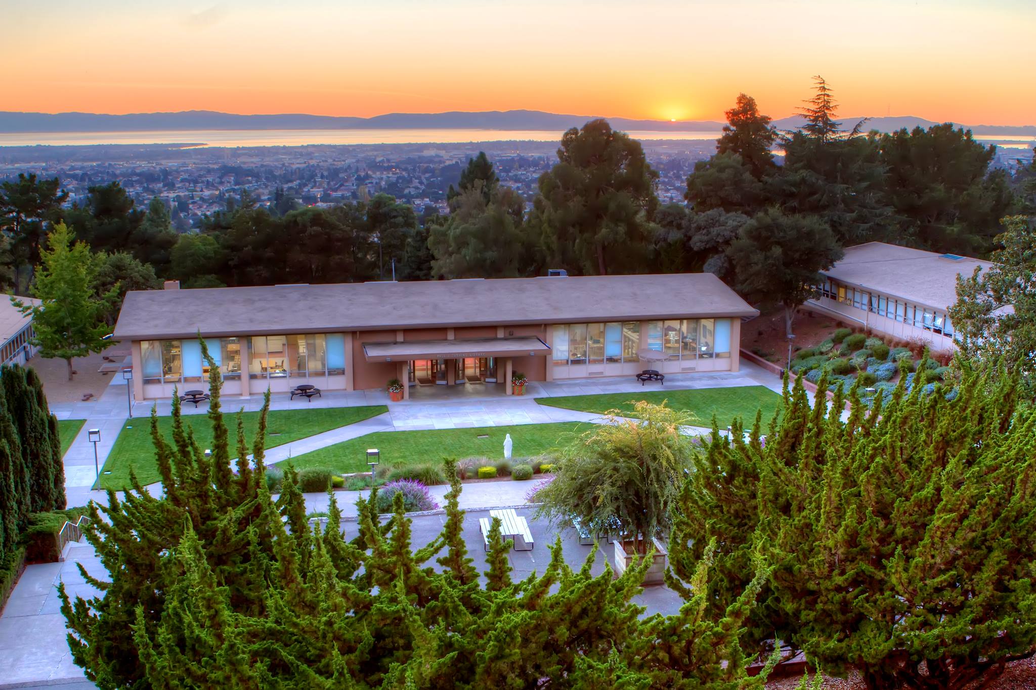 A view of the library at Holy Names University. There is a grassy quad in front of it, with the Oakland skyline in the background at sunset.
